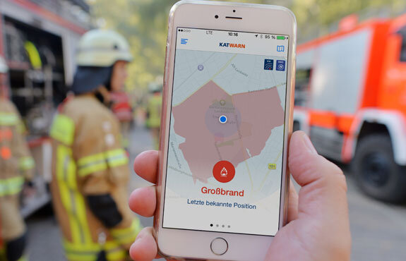 Smartphone zeigt eine Kartenansicht mit der Markierung "Großbrand" im Einsatzfeld, Feuerwehrleute im Hintergrund.